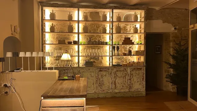 Lit shelves with clay jars and glasses, and a wooden counter facing a stone wall