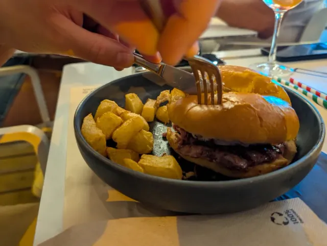 Hands cutting a burger in a dark bowl beside cubes of roasted potatoes