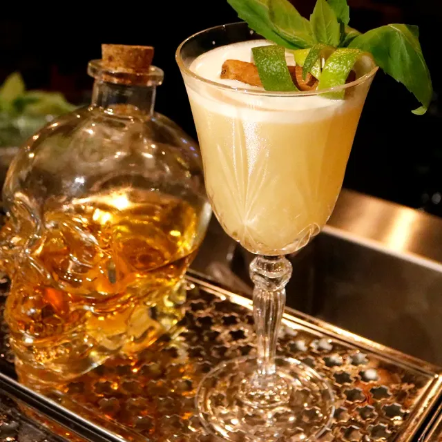 A cocktail in an engraved glass with basil leaves, beside a skull shaped bottle