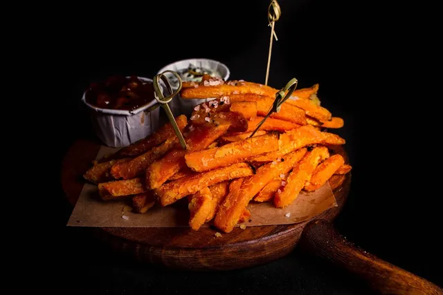 Sweet potato fries on a wooden board with two small bowls of sauce, on a dark background
