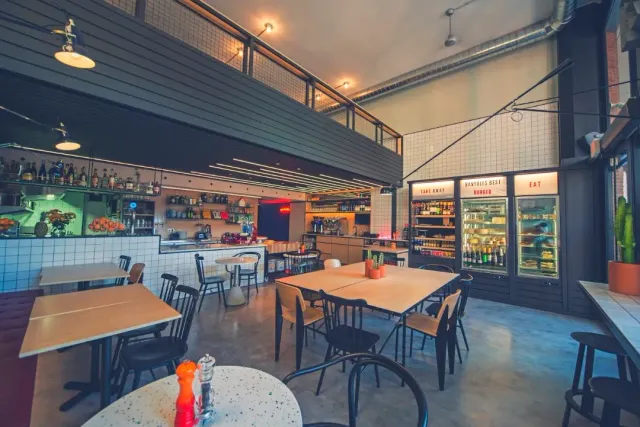 An industrial hall with a white tiled bar, light wooden tables and drink fridges