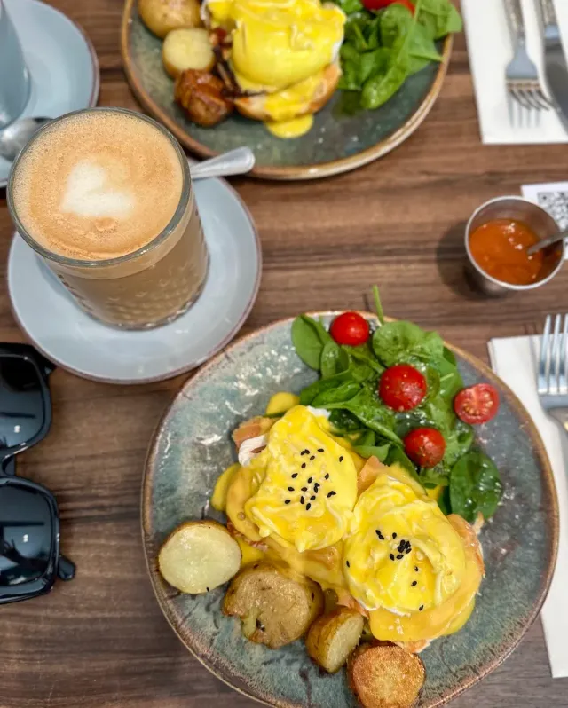 Two plates of eggs in yellow sauce with potatoes and spinach leaves, and a glass of coffee