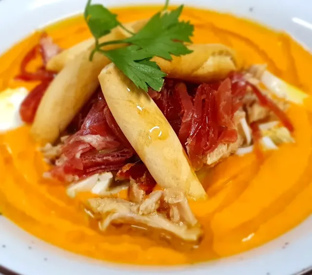 A thick orange soup on a plate with slices of cured meat, bread fingers and green leaves
