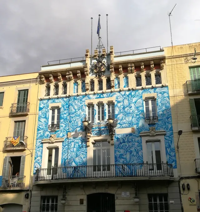 A building facade covered by a blue and white wall painting, iron balconies and flags on the roof