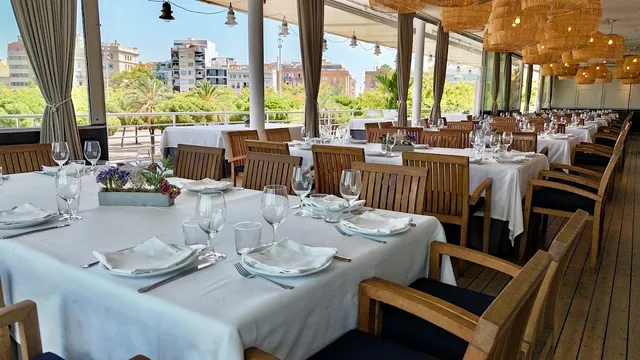 An enclosed terrace with tables set in white cloths and wooden chairs, facing palms and buildings