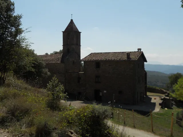 A stone building with a bell tower on a hill, surrounded by trees with hills behind