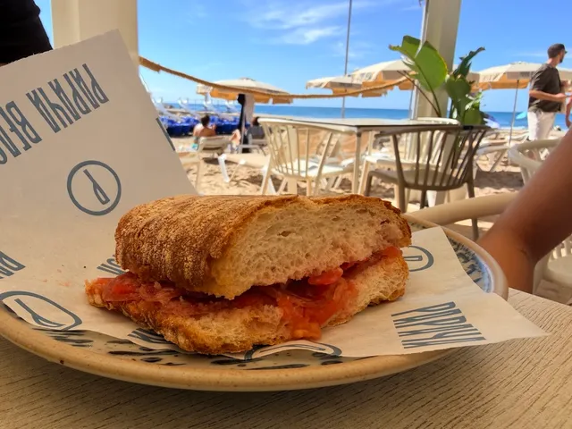A tomato sandwich on wrapping paper, facing a beach with umbrellas and sea