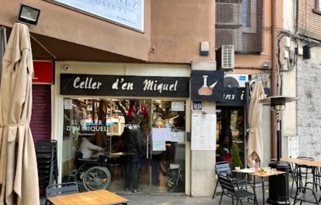 A facade with a black sign and a display window, closed parasols and tables on the pavement