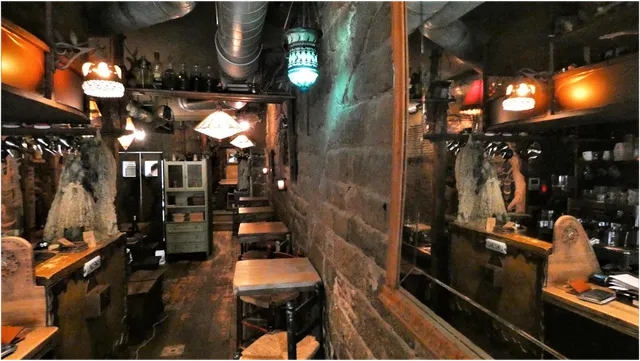 A narrow space with a stone wall, wooden tables and colorful hanging lamps