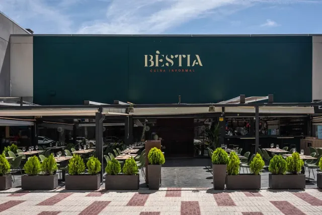 A dark green facade with awnings, outdoor tables and cypress planters along the pavement