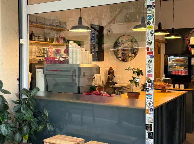 An espresso machine and stacked cups beyond a window, hanging lamps and stickers on the frame