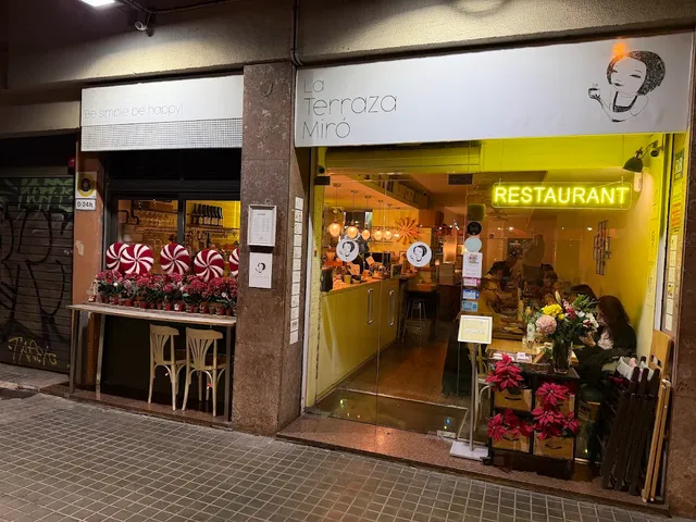 A lit glass frontage in the evening with a yellow neon sign and red flower pots at the entrance