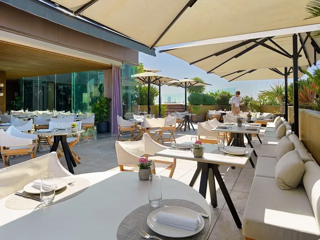 A roof terrace with white parasols, set tables and green plants in sunlight