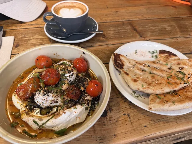 A bowl of labneh with poached eggs and cherry tomatoes beside flatbread and coffee on a wooden table