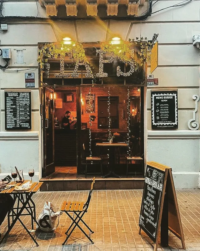 A small bar frontage in the evening with lit letters, chalkboards and a small table on the pavement
