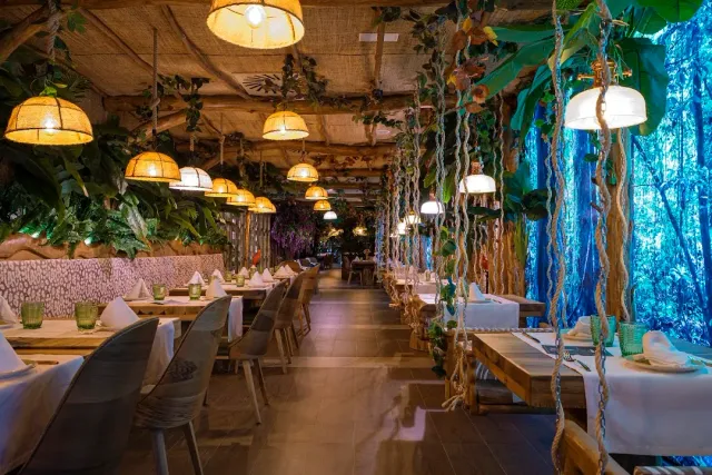 A hall with hanging greenery, straw lamps and long tables on both sides of an aisle