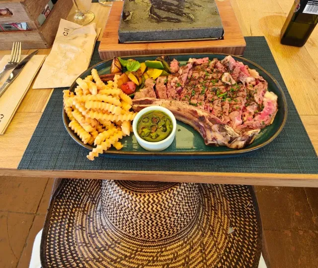 A sliced bone in steak on a plate, with fried potatoes, vegetables and a green sauce