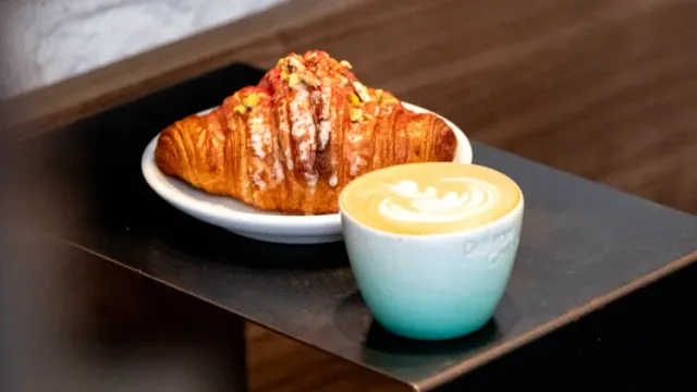 A glazed croissant with pistachio on a white plate, beside coffee in a light blue cup