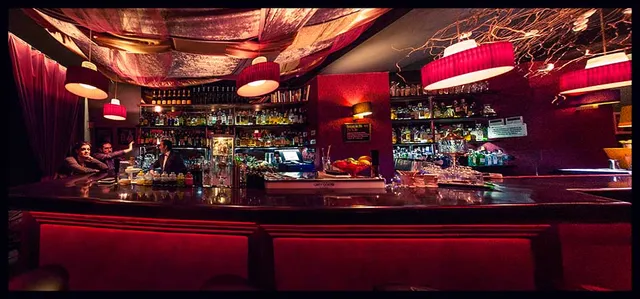 A dark bar counter with red seats, red lamps and lit bottle shelves