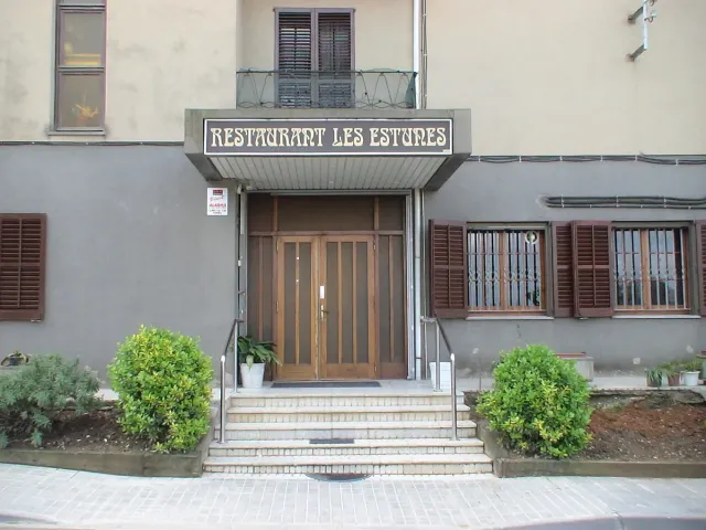 An entrance with a double wooden door, a rectangular sign above it and steps between shrubs