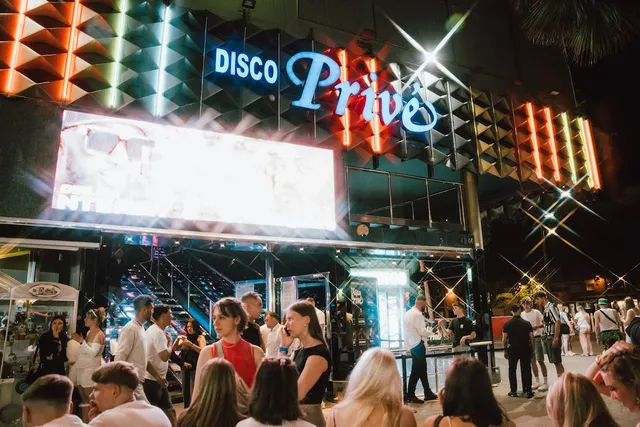 A crowd of young people at night in front of a club facade with a neon sign and a large screen