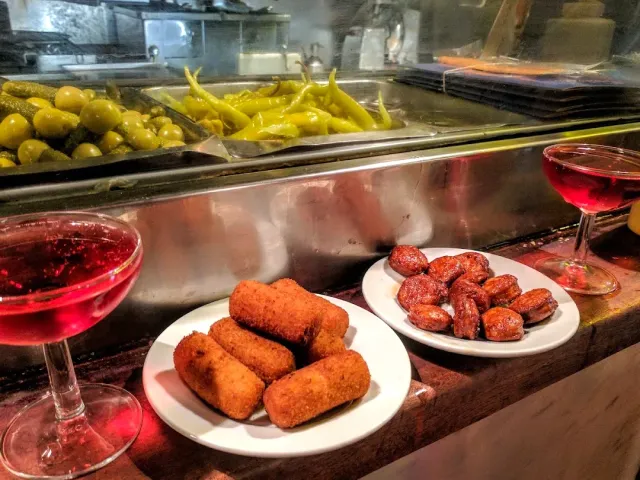 A plate of fried dough rolls and a plate of red sausages, beside two glasses of red drink