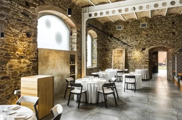 A hall with rough stone walls and arches, round tables in white and dark chairs