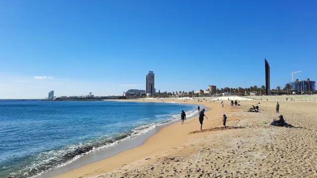 A sandy stretch of beach facing a blue sea, a few walkers and city towers on the horizon