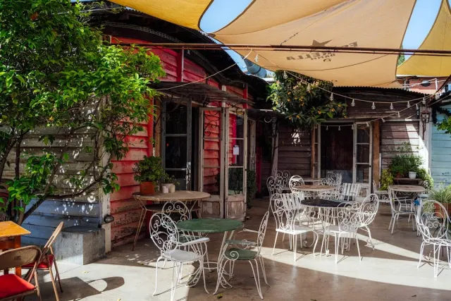 A shaded courtyard with white iron chairs, a colorful wooden cabin and a string of lights