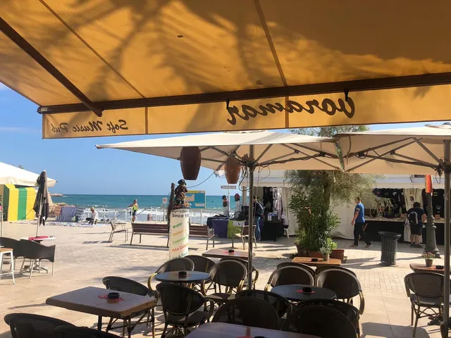 A terrace with tables in the shade of a yellow awning, facing a promenade and blue sea