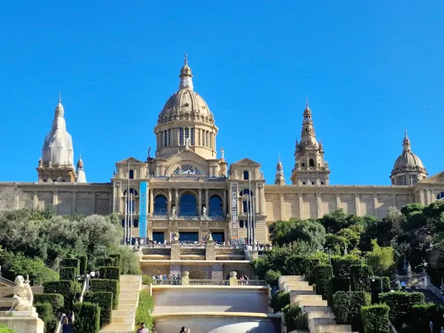 A wide stone palace with a dome and towers, steps and a fountain in front of it