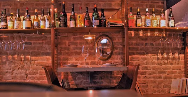 Shelves of wine bottles above a small table by an exposed brick wall and a round mirror