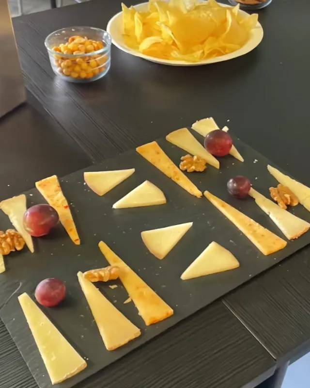 A dark board with cheese wedges, grapes and walnuts, beside a small bowl of corn and a plate of crisps
