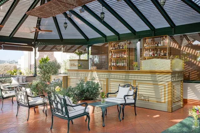 A bar under a glass roof with green iron chairs, potted plants and a terracotta floor