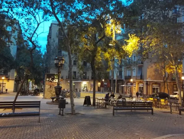 A paved square with trees, an iron lamp post, benches and terrace tables in the evening