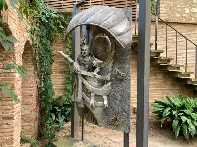 A metal sculpture of a figure in a ship beneath a large leaf, in a courtyard with brick arches