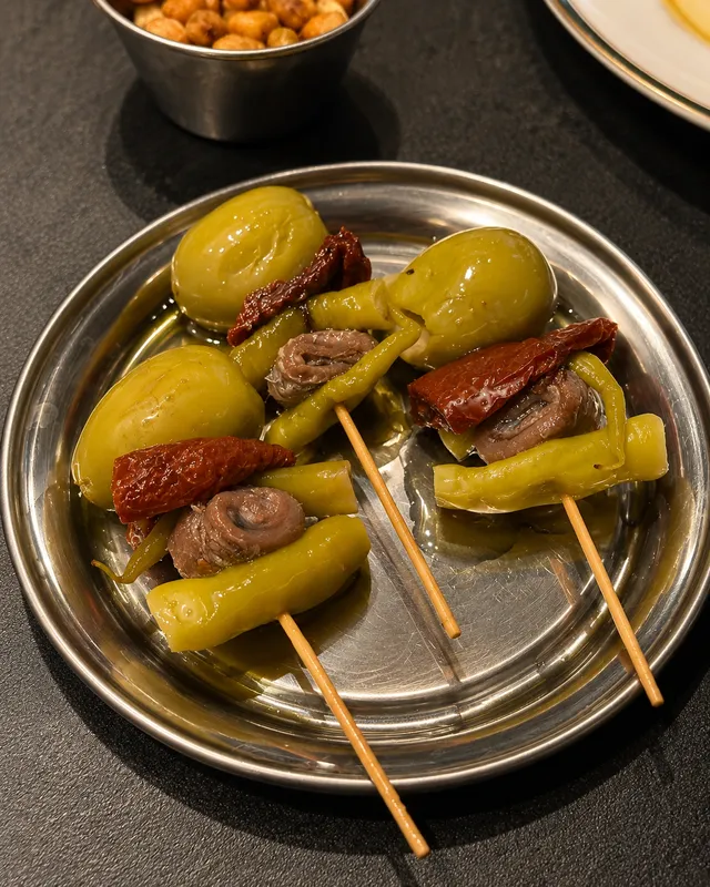 Skewers of olives, green peppers and anchovies on a metal plate, beside a bowl of snacks