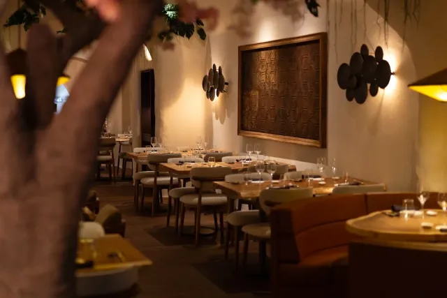 A softly lit hall with wooden tables, upholstered chairs and a tree trunk in the foreground