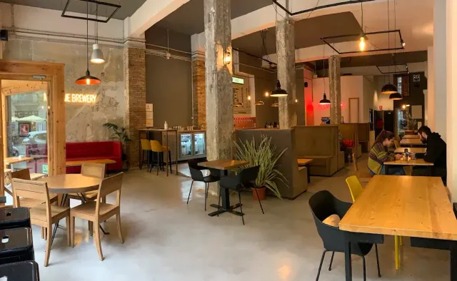 A hall with concrete columns and an exposed brick wall, wooden tables and a red sofa