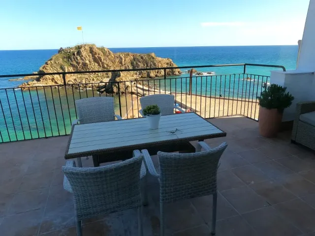 A table and four wicker chairs on a terrace facing blue sea and a rocky cliff