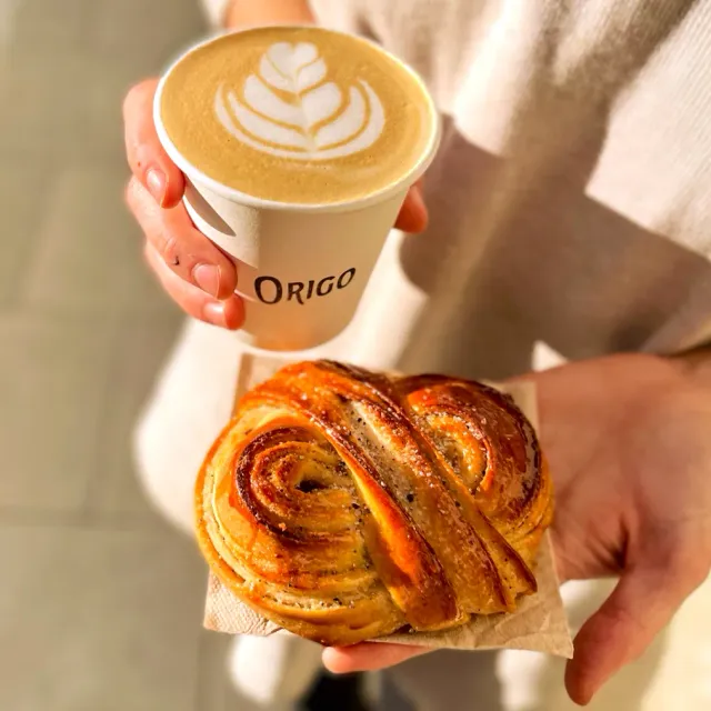 A hand holding a coffee cup with foam art, and a second hand holding a swirled pastry
