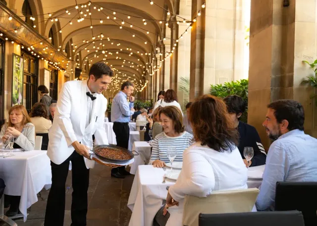 A waiter in a white jacket serving a rice pan to a table under stone arches