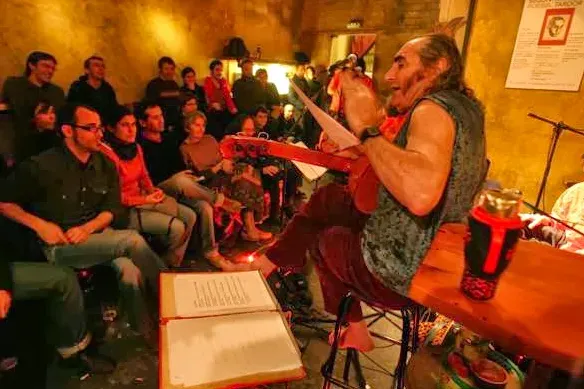 A musician playing a string instrument before a packed audience in a small room in reddish light