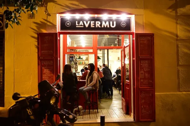 A lit entrance with a sign above, chalkboards at the sides and people sitting inside in the evening