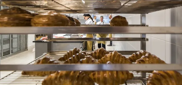 Croissants on metal racks in front, and bakers working in the kitchen behind