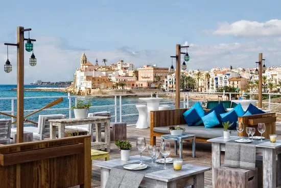 A wooden terrace with sofas and blue cushions, facing a bay and a seaside town with a church
