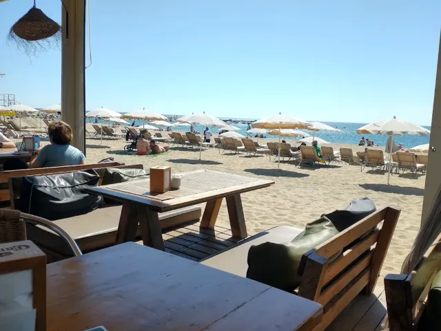 A wooden table in the shade facing a beach with parasols, sun loungers and blue sea