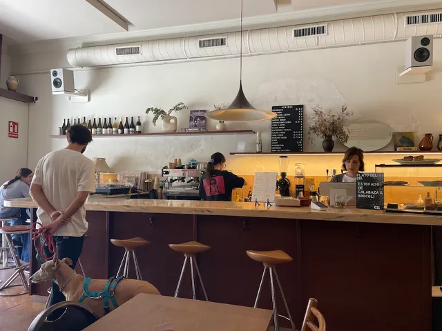 A long wooden counter with stools and a shelf of wine bottles, and diners in a bright room
