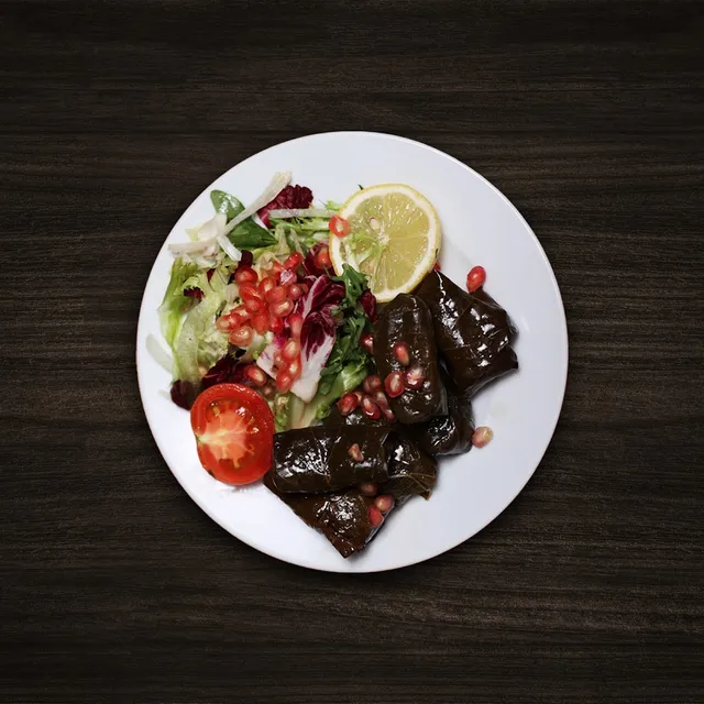 Stuffed vine leaves beside a green salad, tomato, lemon and pomegranate seeds on a white plate
