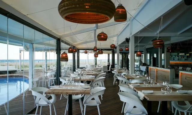 A bright dining room with white chairs and straw lamps, a pool and sea beyond the glass